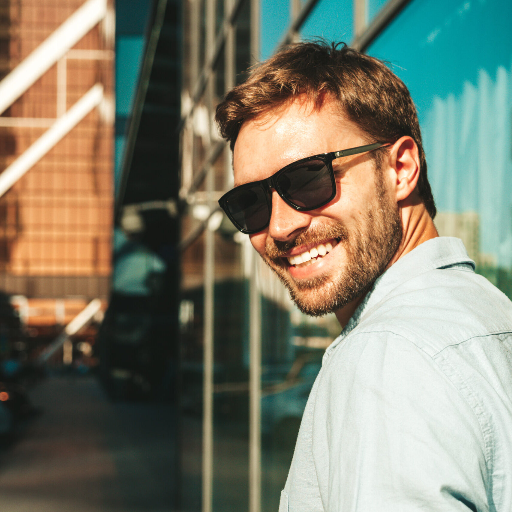 Portrait of handsome smiling stylish hipster lambersexual model.Modern man dressed in blue shirt. Fashion male posing in the street background near skyscrapers in sunglasses